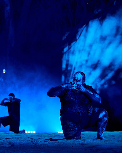 Two Noongar men perform a dance. They are wearing neon paint on their faces and the lighting is blue. It is nightime.